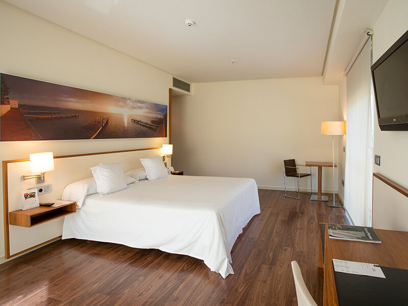 Habitación de hotel con cama grande, mesitas de noche, escritorio, silla y TV. Arte paisajístico sobre la cama.