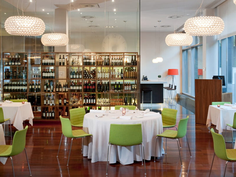Interior de restaurante moderno con estanterías de vino, mesas y sillas verdes.