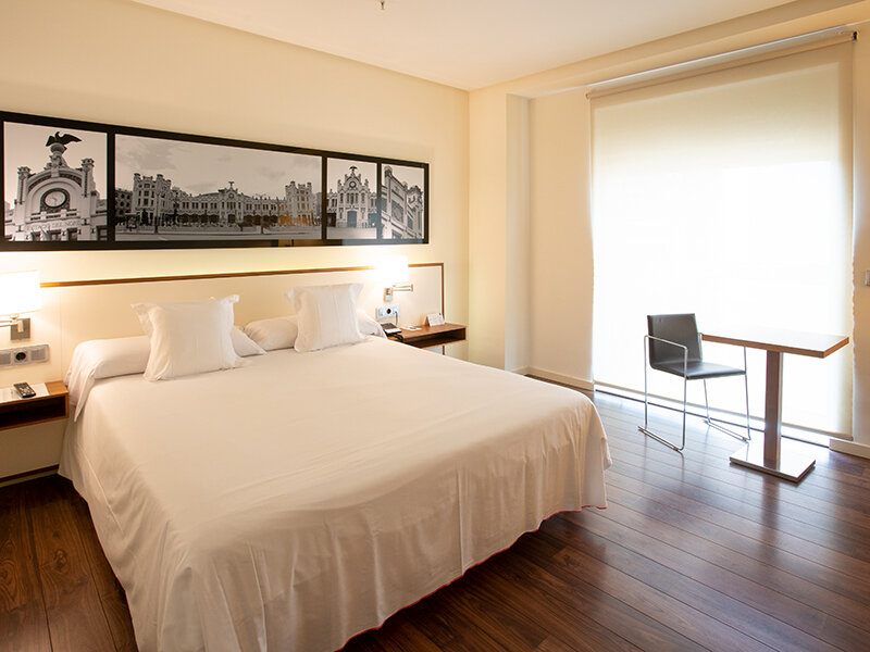 Una habitación de hotel con cama blanca, mesita de noche, cuadro y silla en una mesa.