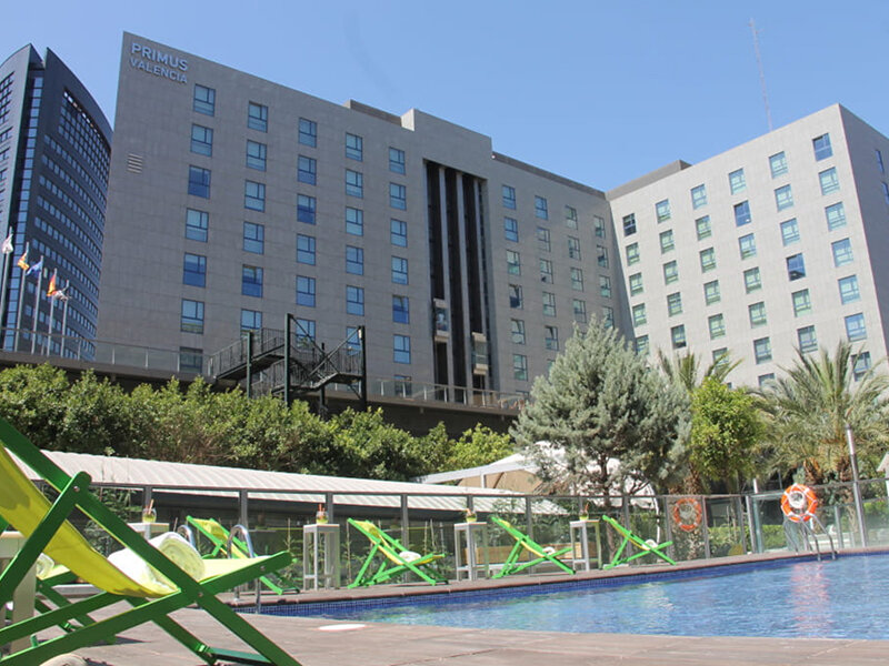 Tumbonas verdes junto a la piscina frente al hotel Primus Valencia con árboles y otro edificio.