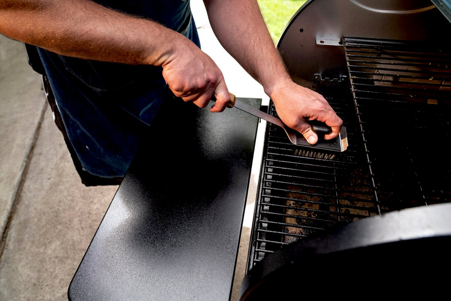 Hombre limpia parrilla con cepillo junto a mesa negra.