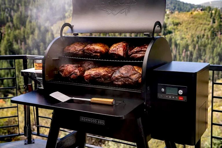 Una parrilla con carne en dos estantes, humo ascendente, al aire libre con árboles al fondo.