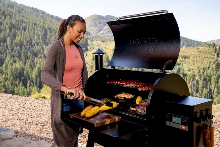 Una mujer asa carne y maíz al aire libre con montañas de fondo.