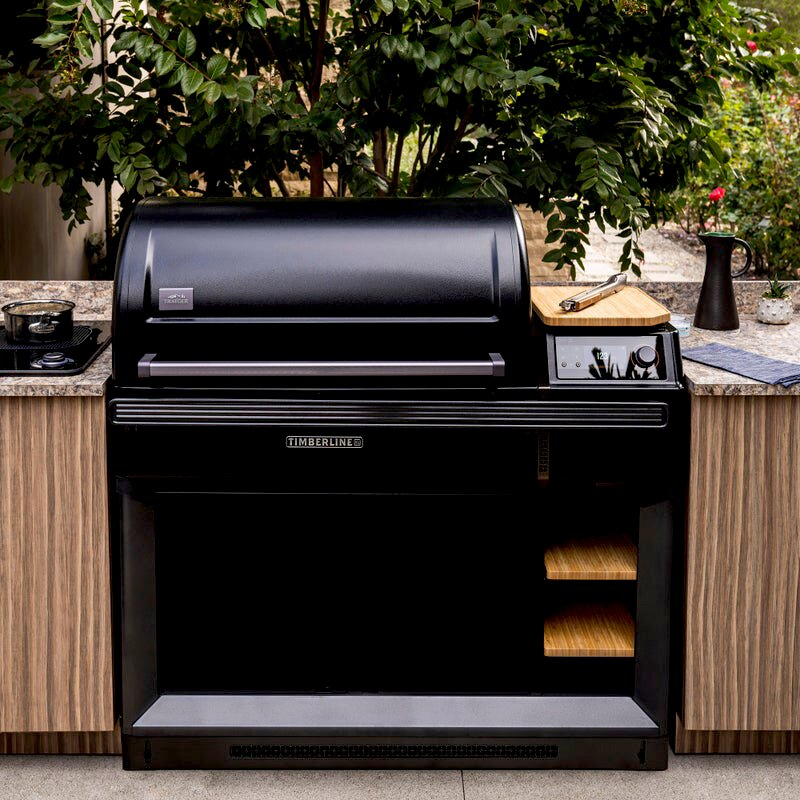 Una parrilla Timberline negra está al aire libre, con vegetación de fondo y una encimera.