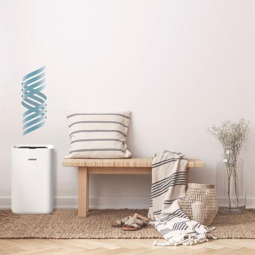 A white air purifier sits beside a wooden bench with cushions, blankets, and a basket.