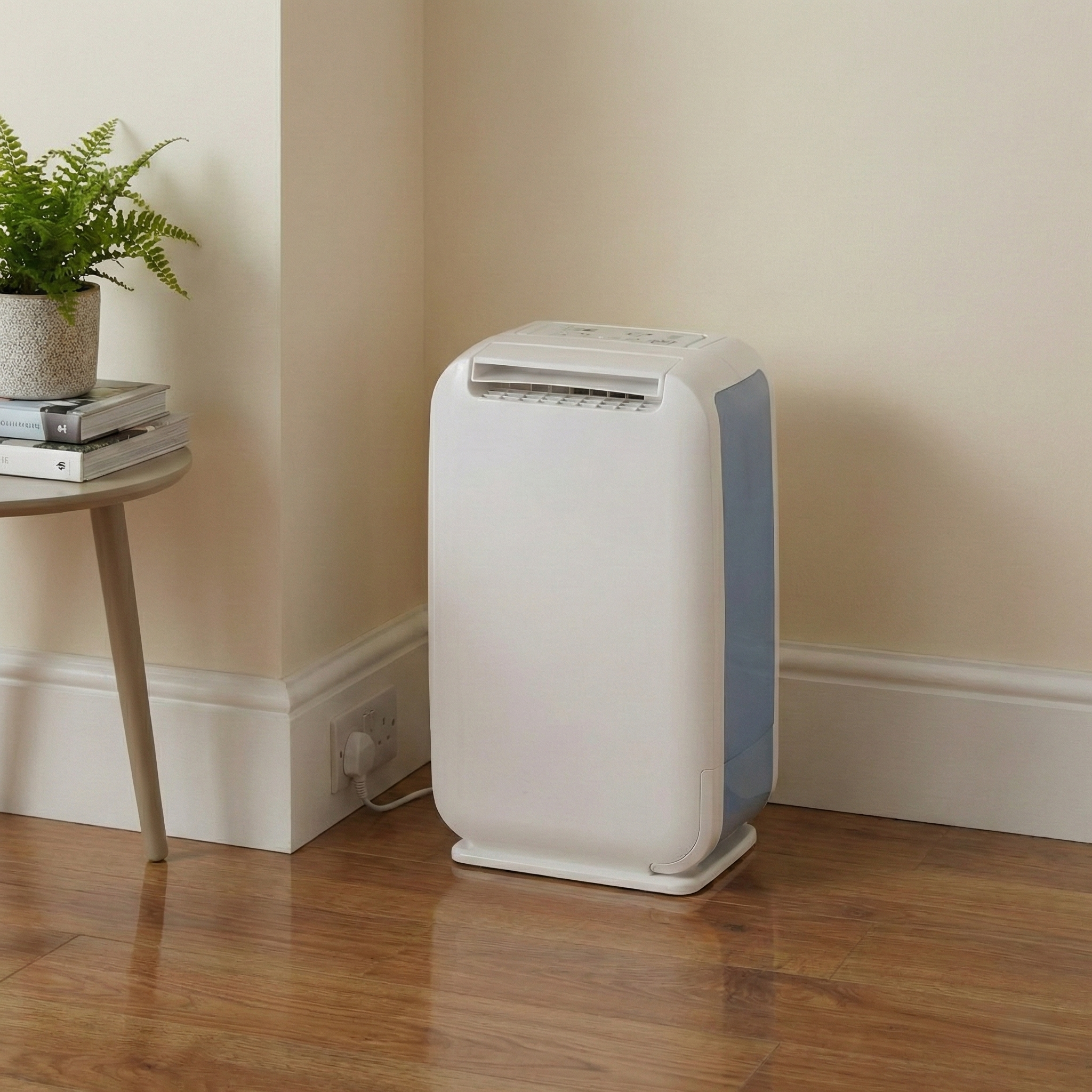 A white dehumidifier with blue accents stands beside a table with books and a plant.