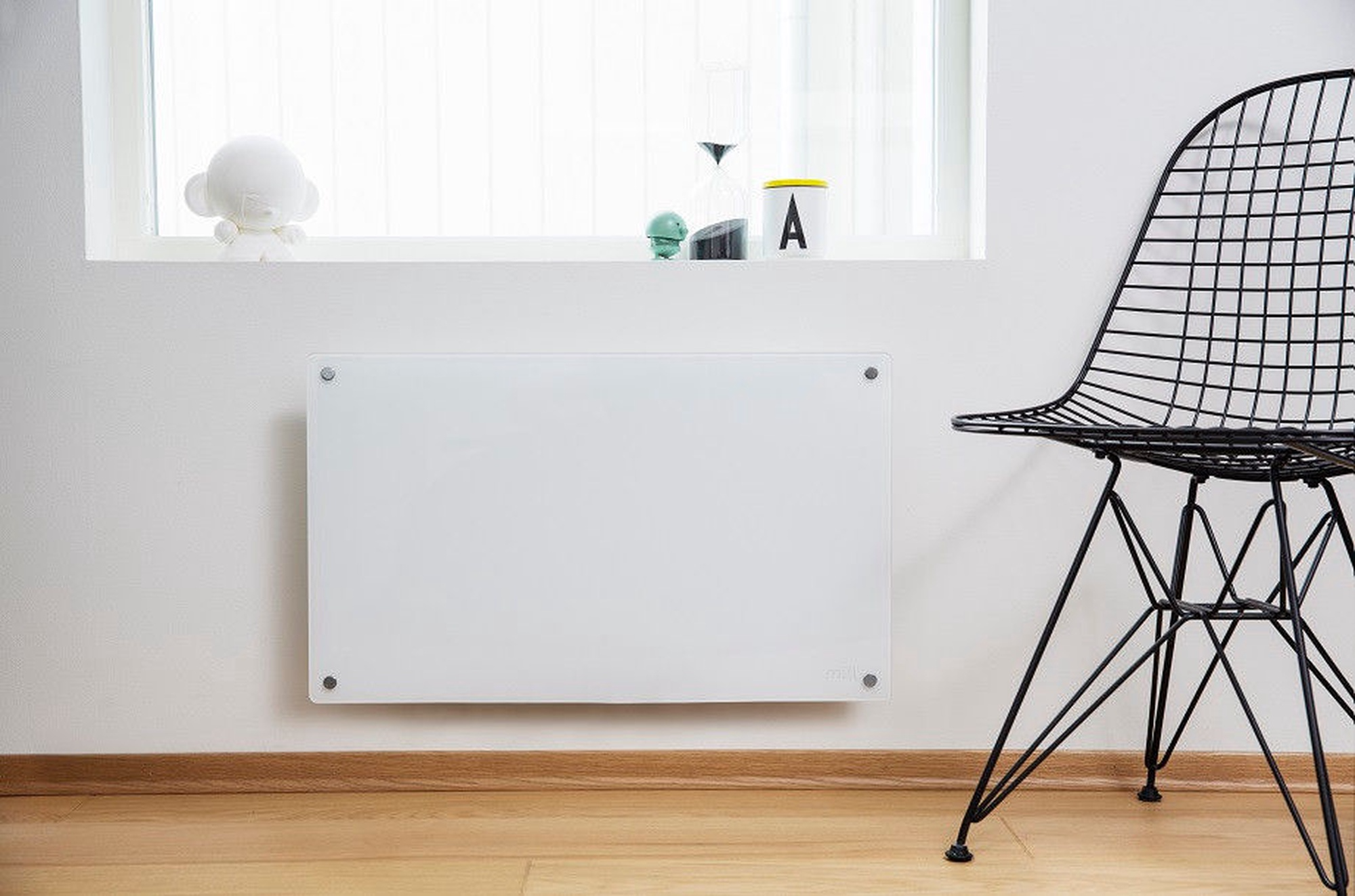 A white glass heater on a wall with a wire chair and window sill decor.