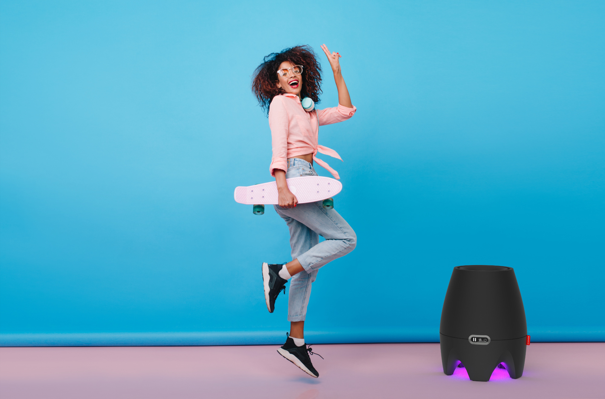 A woman with curly hair holds a pink skateboard next to a black speaker.