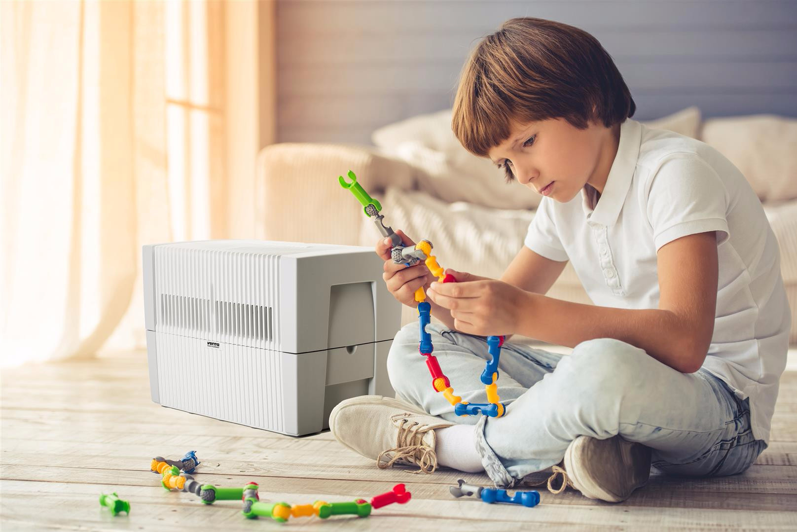 A boy plays with colorful building blocks next to a white air purifier on a wooden floor.