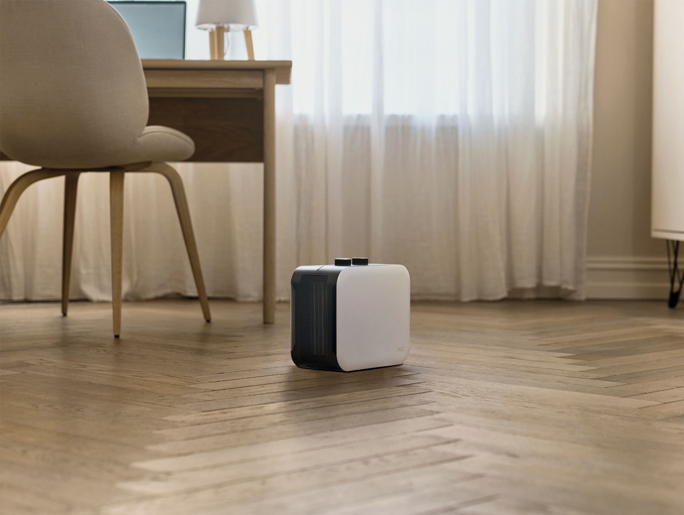 A white heater sits on a wooden floor in front of a window with curtains.