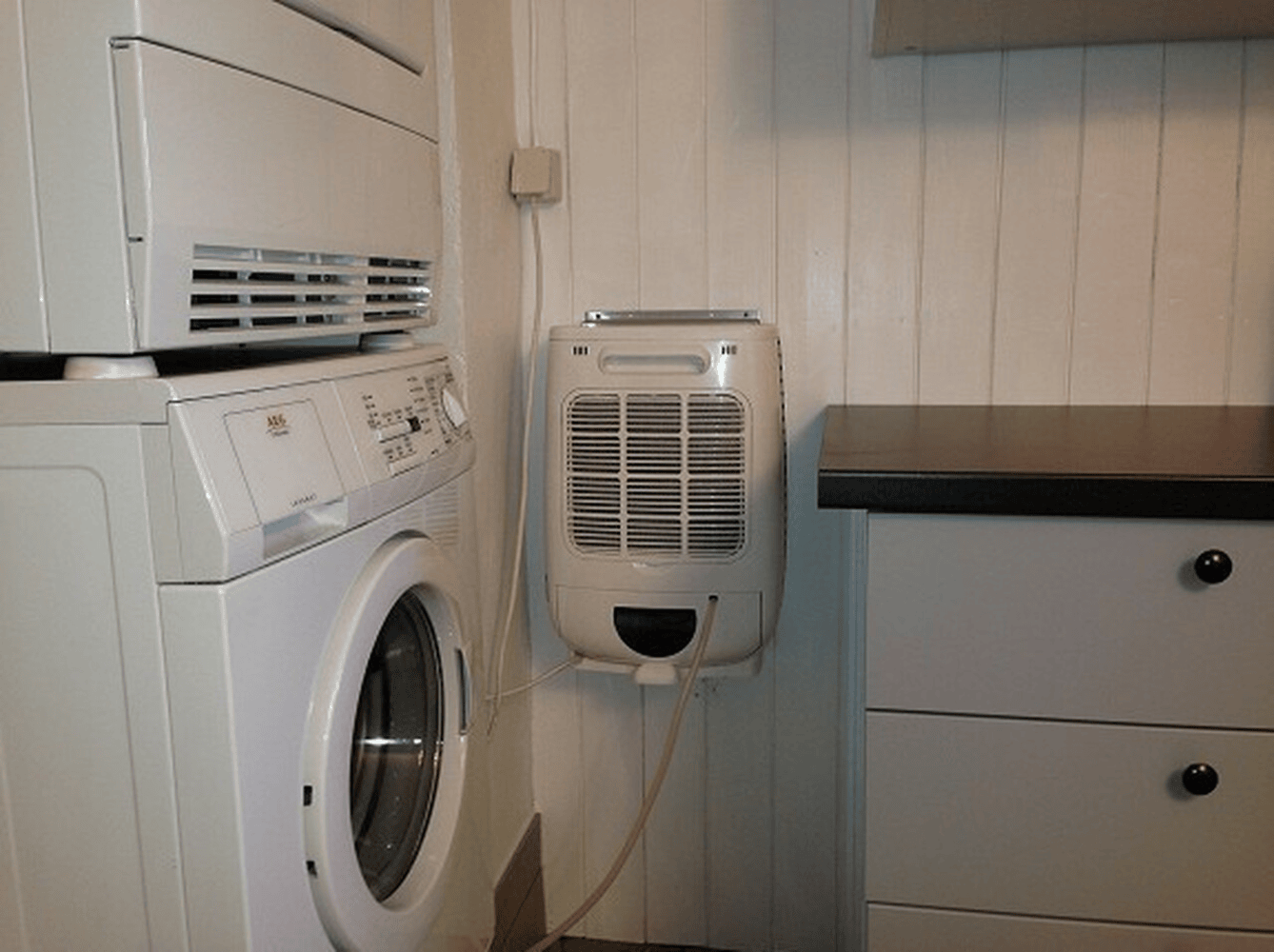 A white washing machine and dryer are stacked. A white dehumidifier is on the wall.