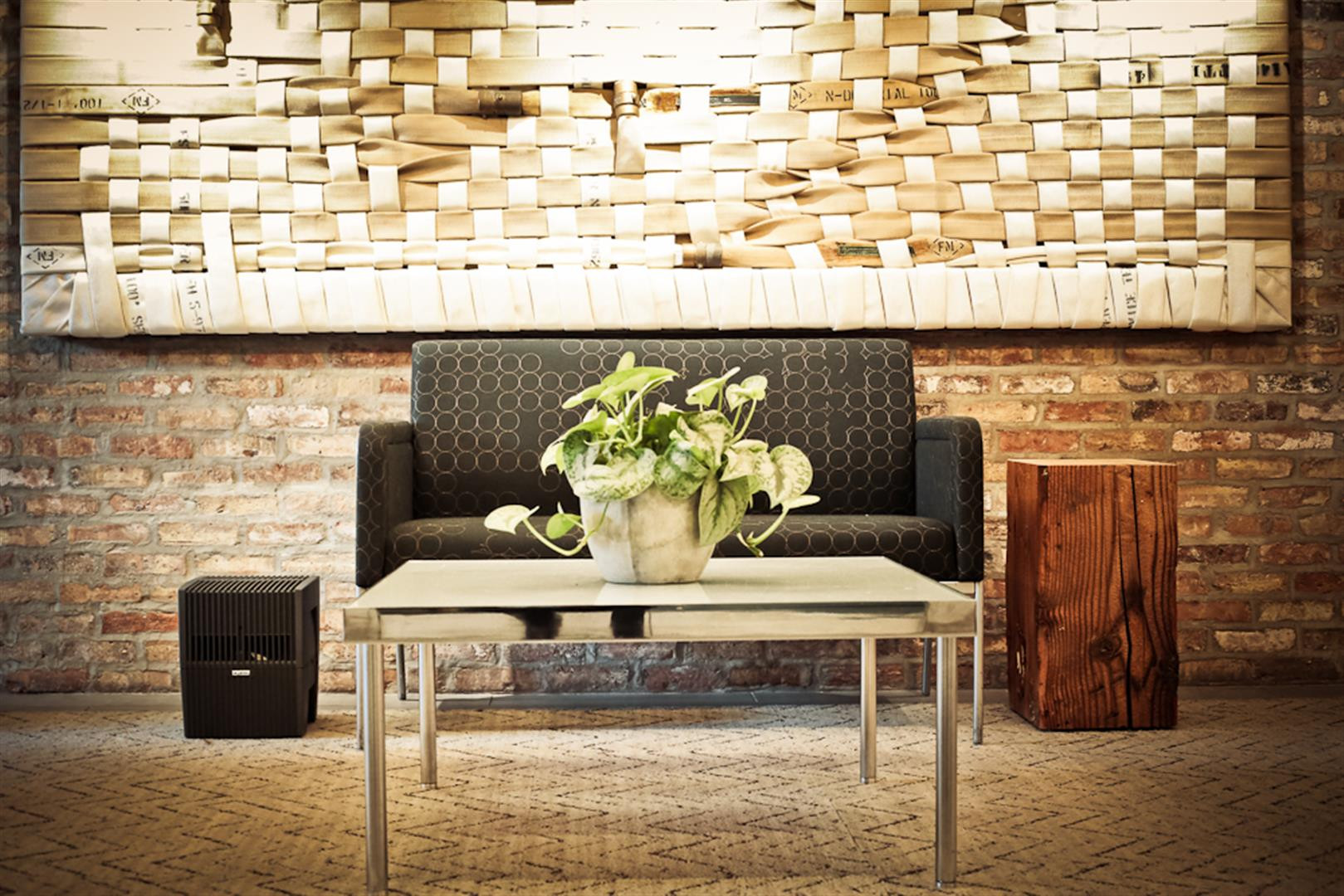 A sofa with a plant sits before a brick wall and a tapestry.
