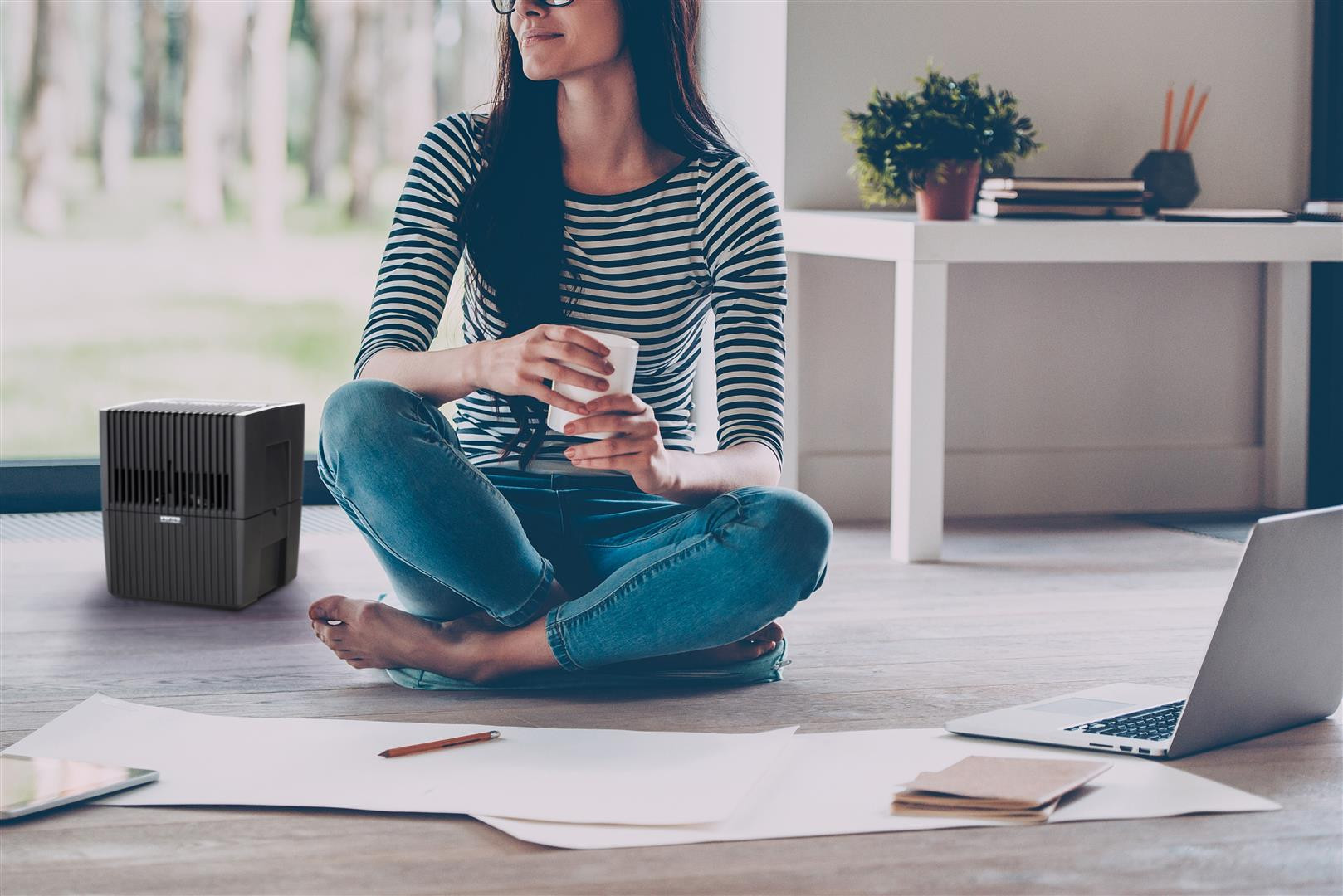 A woman sits on the floor beside an air purifier, holding a cup.