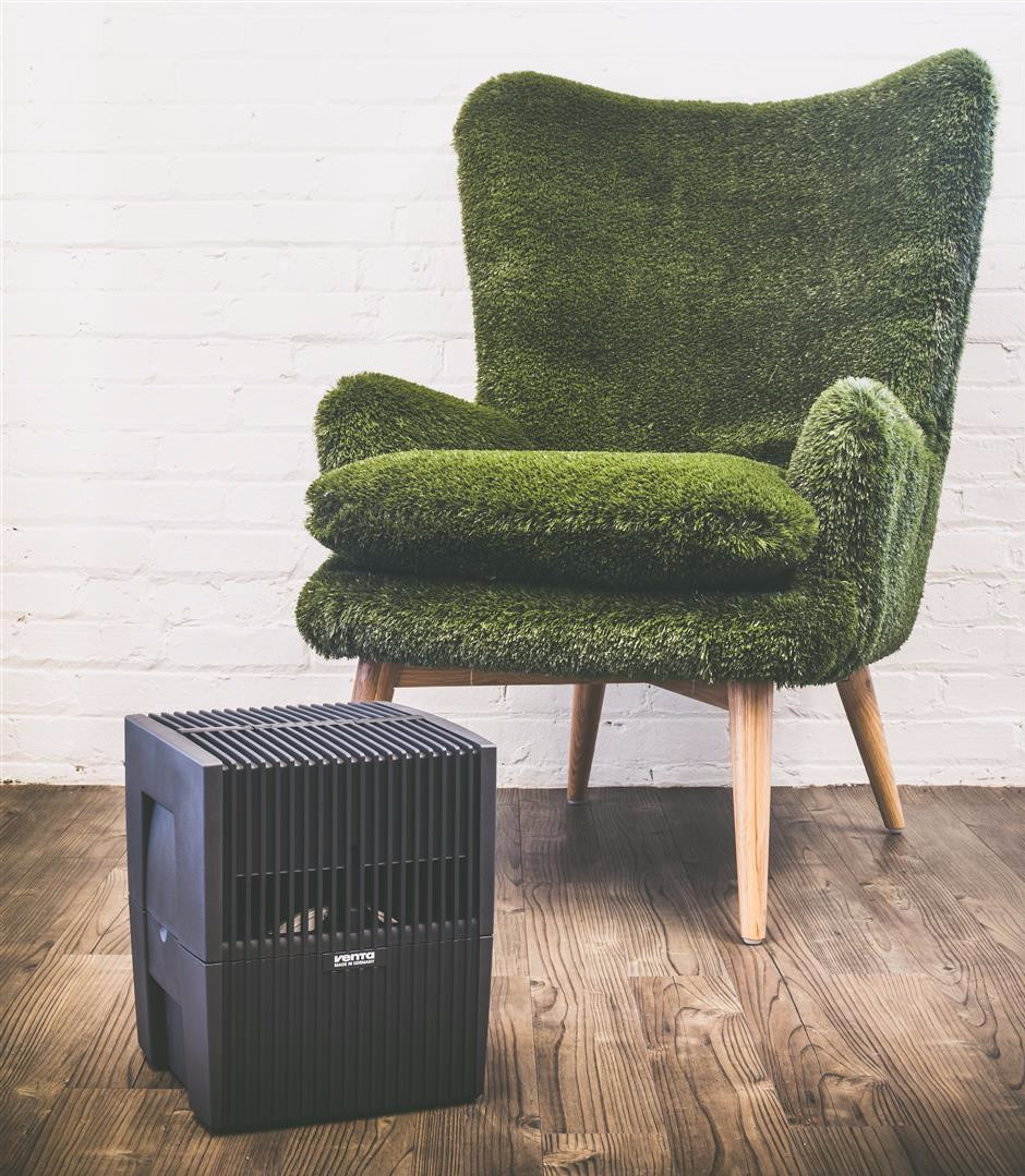 A black air purifier sits on a wooden floor next to a green grass-covered armchair.