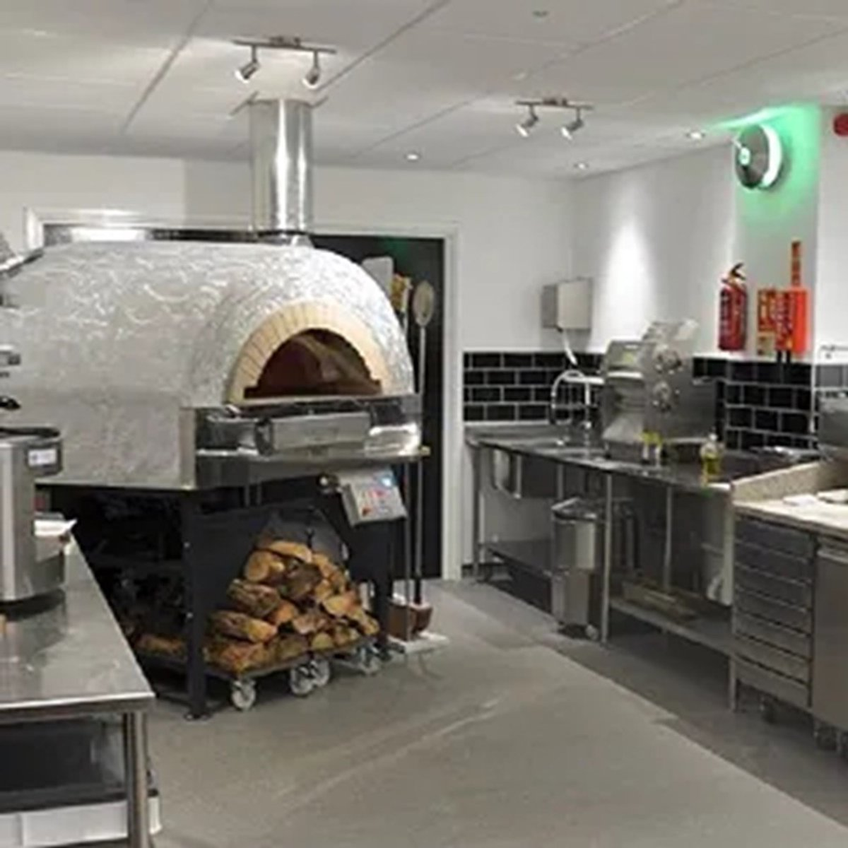 A pizza oven with firewood, stainless steel counters, and equipment in a kitchen.