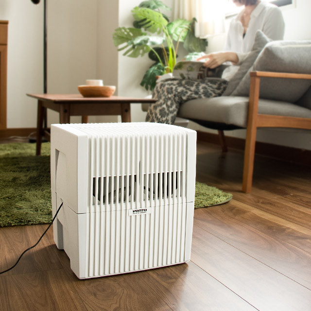 A white humidifier sits on a wooden floor in front of a sofa where a woman reads.