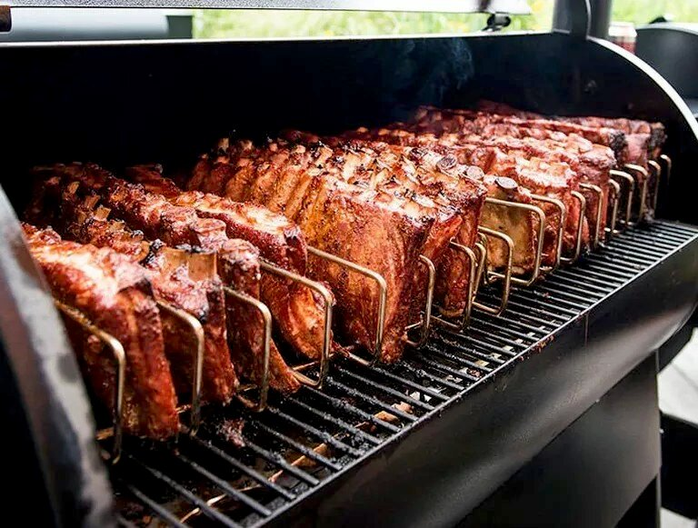 Filas de costillas cocinándose en una parrilla, sujetas en soportes metálicos. Humo.