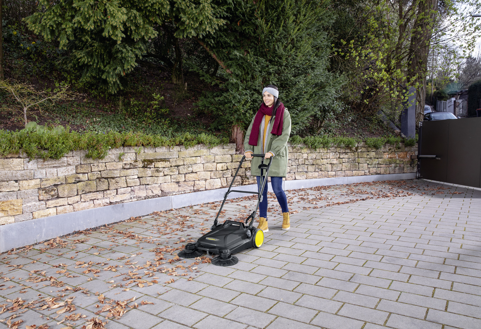 Una donna spinge una macchina spazzatrice su un vialetto pavimentato con foglie cadute.