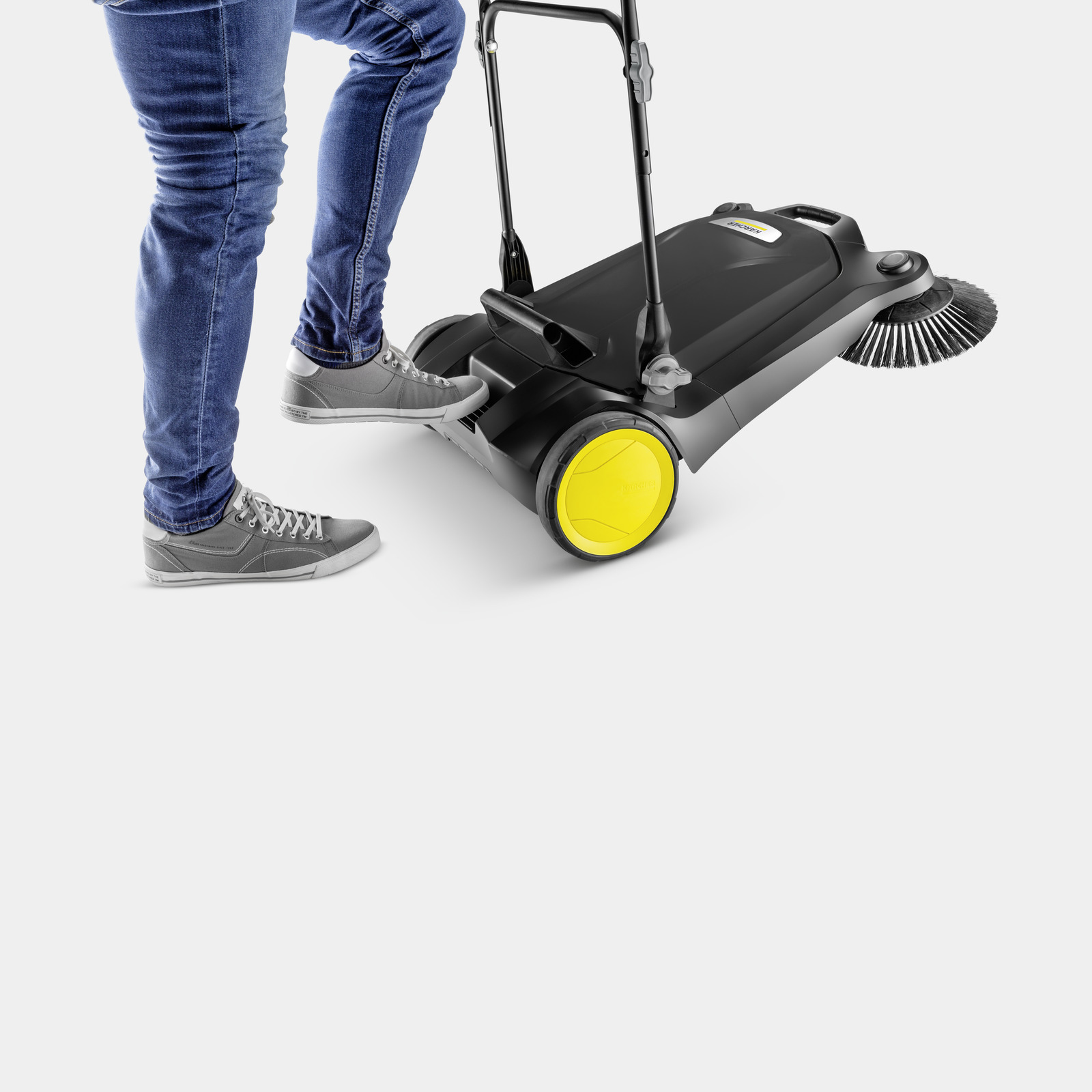 Persona in jeans e scarpe da ginnastica usa una scopatrice a spinta su sfondo bianco.