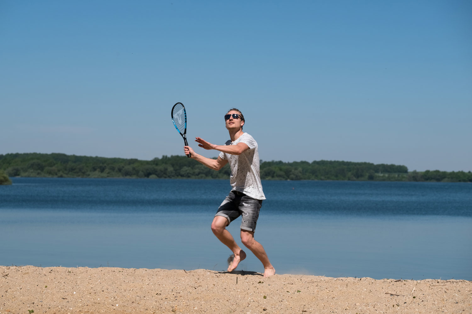 Un uomo gioca a tennis su una spiaggia con un lago sullo sfondo.