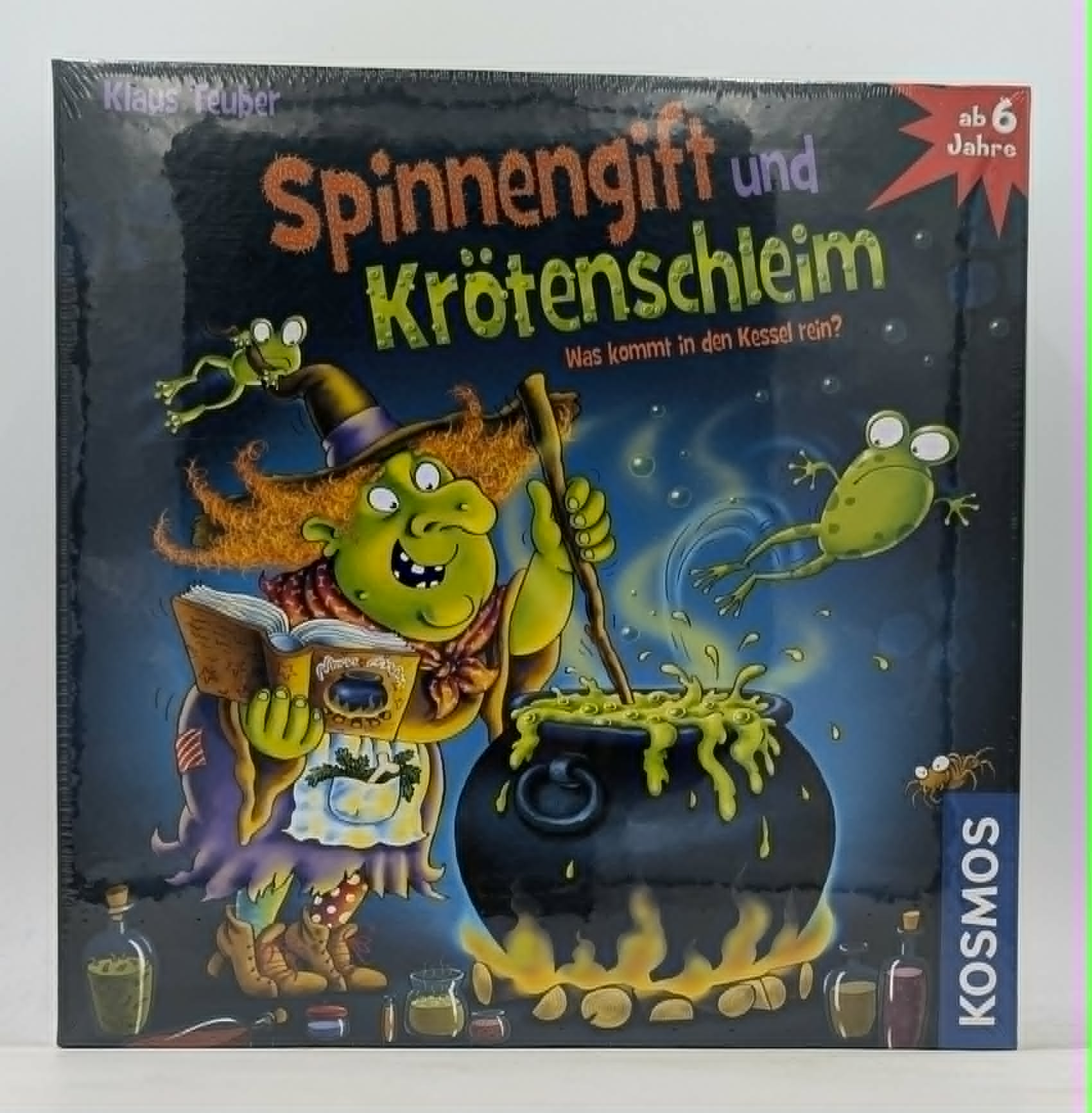 Eine grüne Hexe kocht in einem Kessel, Frösche springen. Spielverpackung.