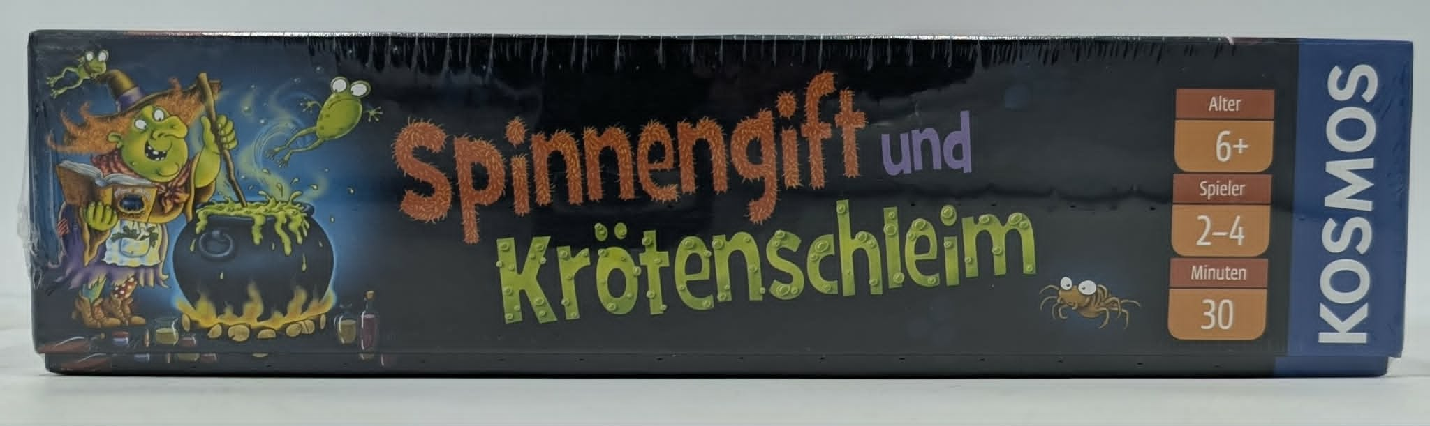 Ein grüner Hexen-Spielkarton mit Fröschen, Spinnen, Kessel und Text.