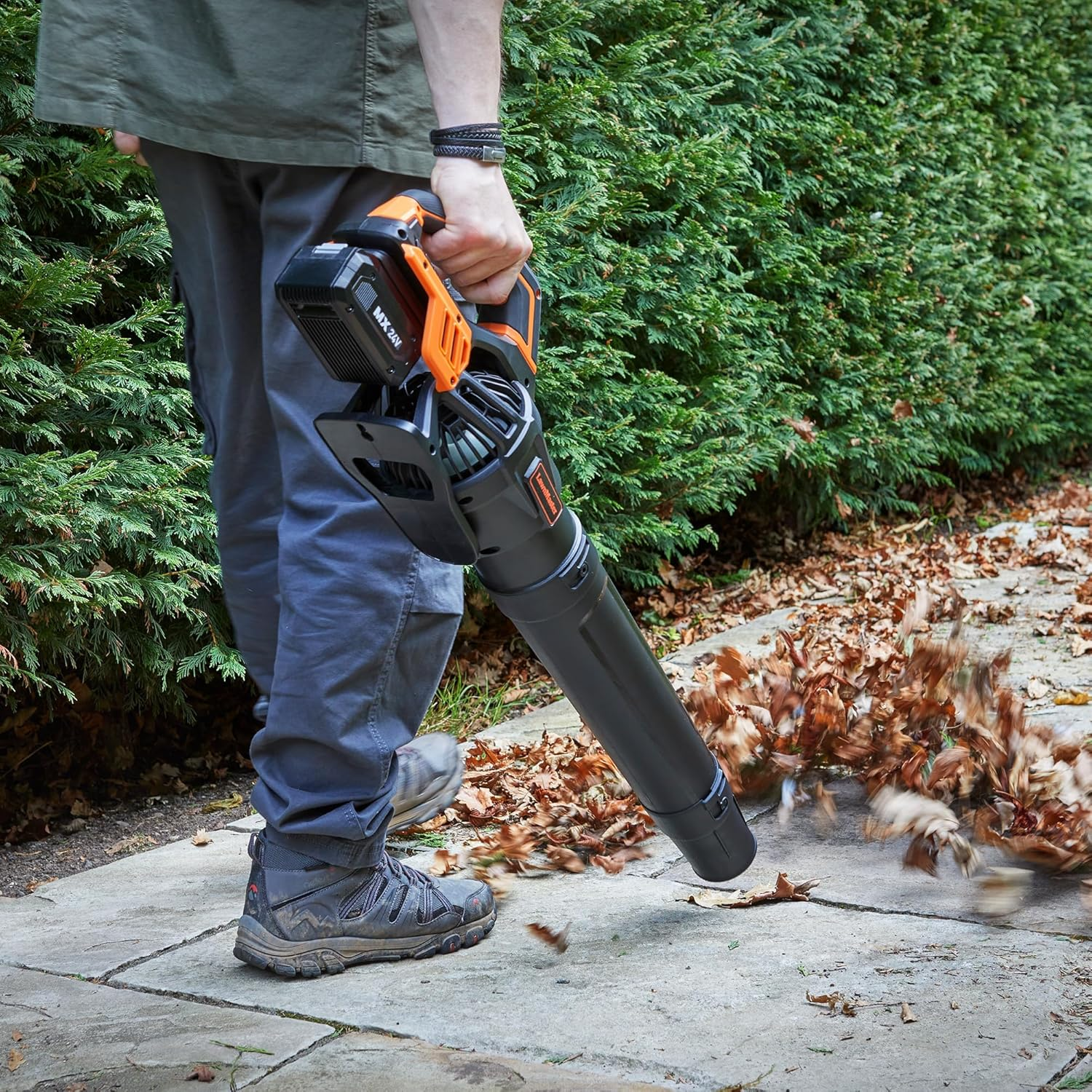 Person fegt mit Laubbläser trockene Blätter von einem Gehweg im Freien.