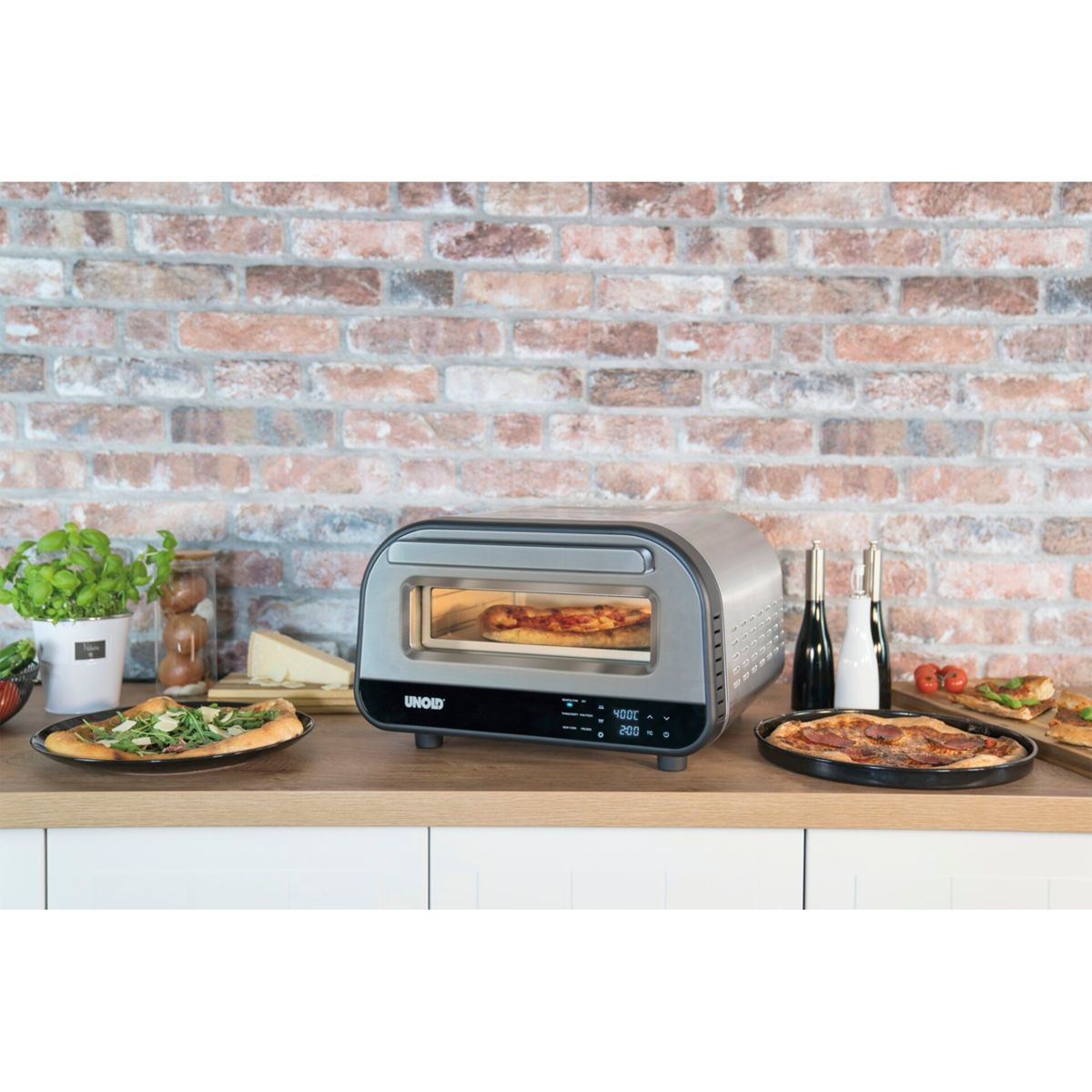 Ein silberner Toaster backt eine Pizza. Zwei Pizzen, Käse und Basilikum stehen auf der Theke.