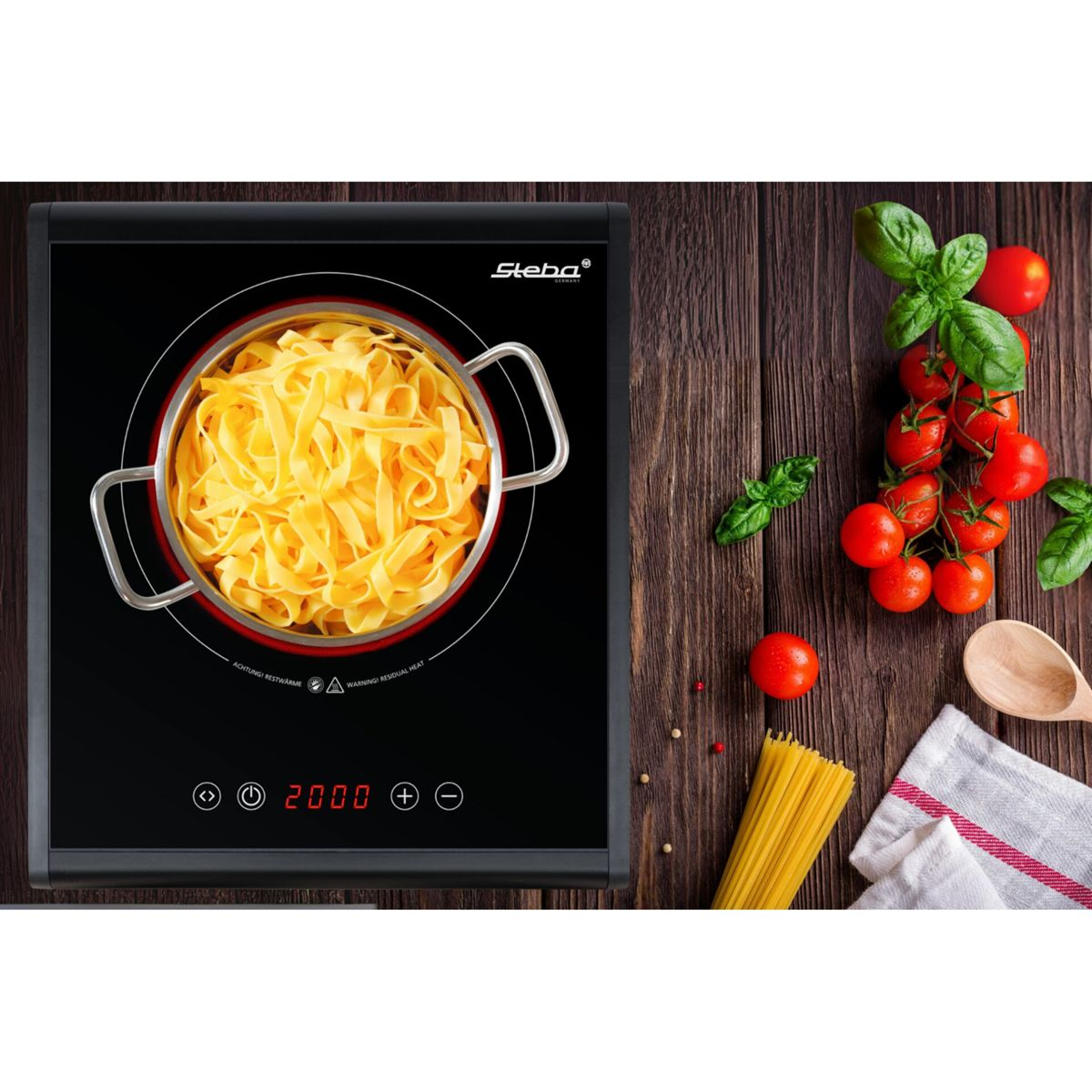Una pentola di fettuccine su un piano cottura a induzione, con pomodori e basilico nelle vicinanze.