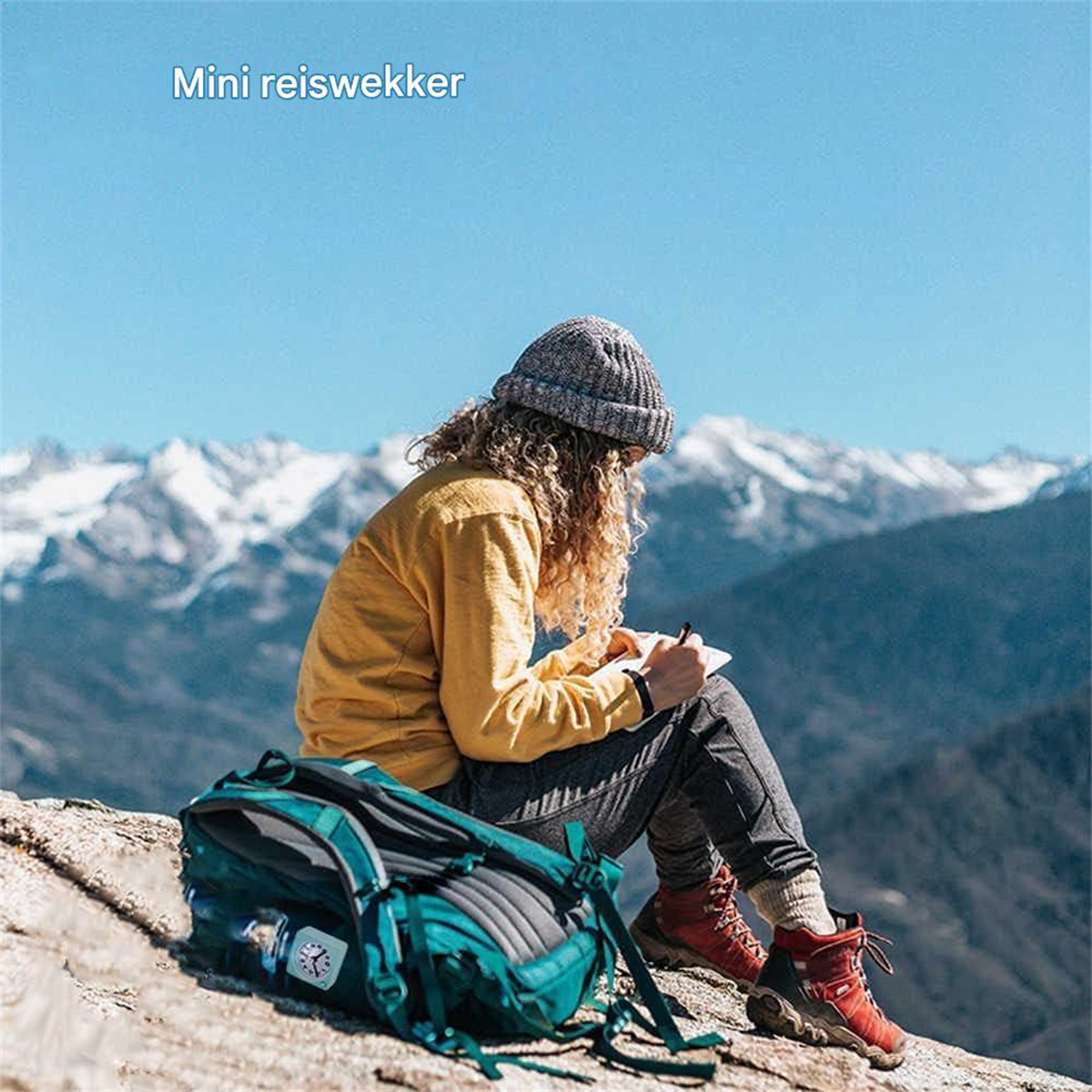 Eine Frau mit lockigem Haar sitzt auf einem Felsen und schreibt in ein Notizbuch. Sie trägt eine gelbe Jacke und eine graue Mütze.