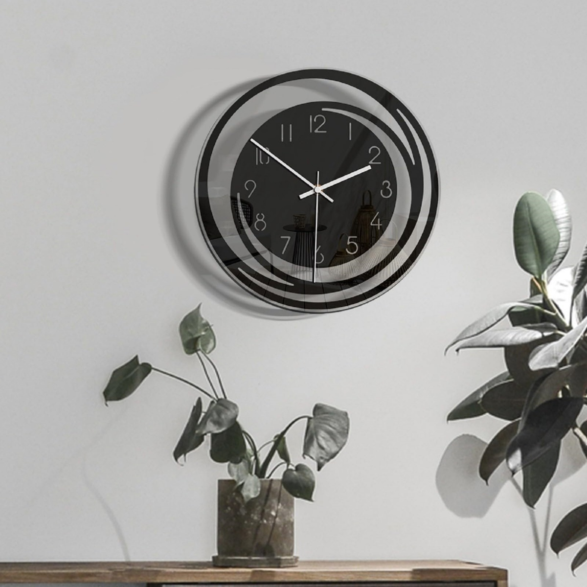 Eine schwarze Wanduhr mit weißen Zeigern hängt an einer weißen Wand. Links davon steht eine Pflanze.