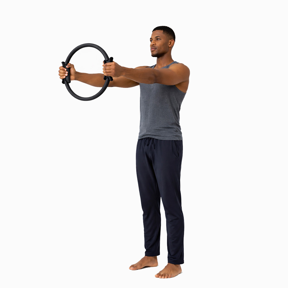 Un uomo con canottiera grigia e pantaloni scuri tiene un anello Pilates nero a braccia tese.