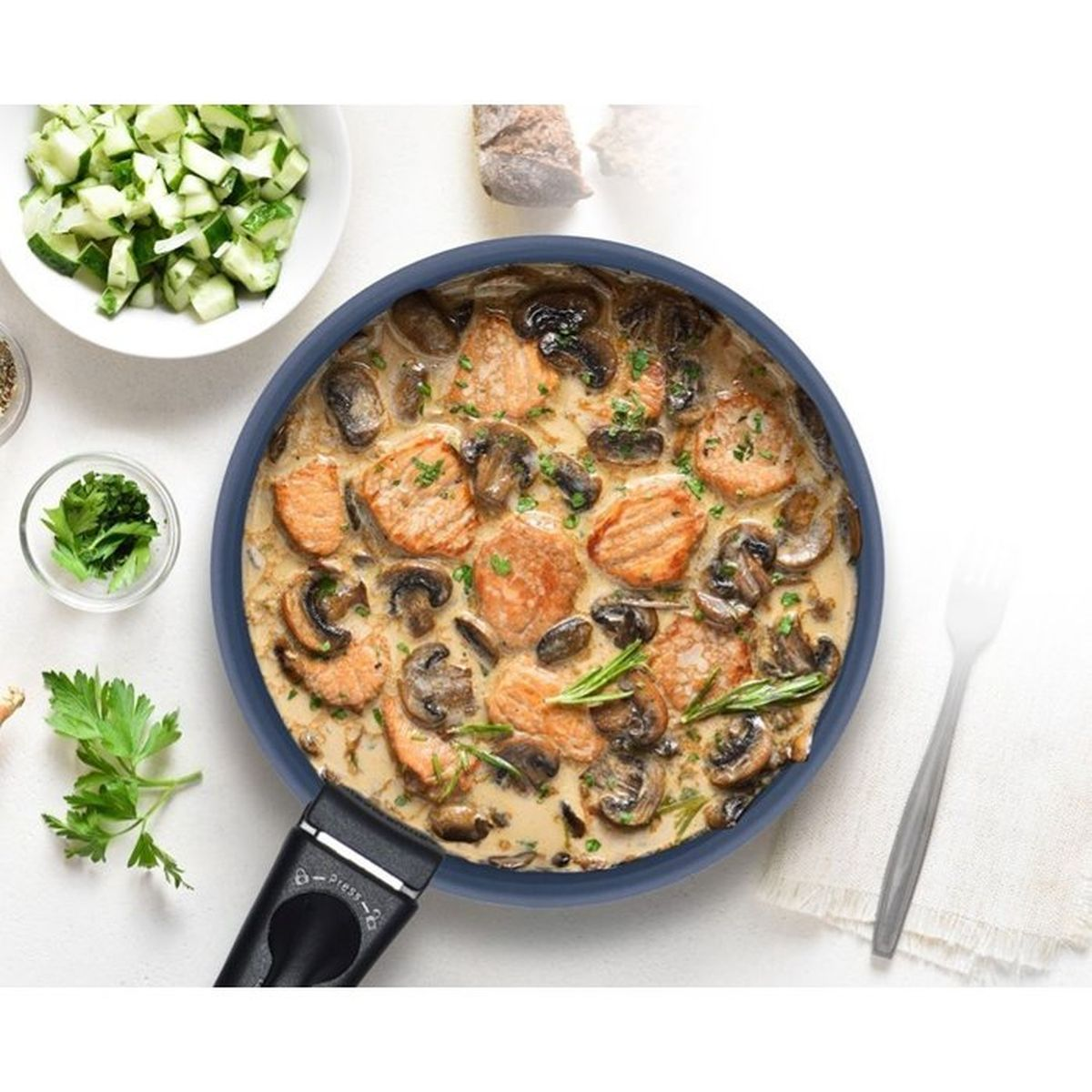 Fleisch in cremiger Pilzsauce in einer Pfanne, daneben Gurkensalat und Kräuter.