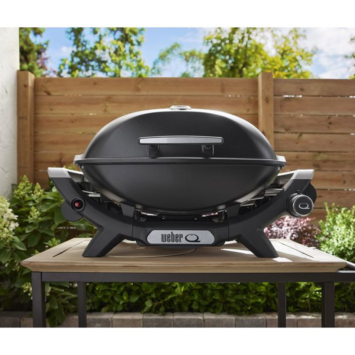Ein schwarzer Weber Q Grill steht draußen auf einem Holztisch, im Hintergrund ein Holzzaun und Grünzeug.