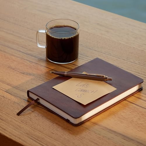 Eine gläserne Tasse Kaffee steht neben einem braunen Notizbuch und Stift auf einem Holztisch.
