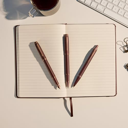 Drei roségoldene Stifte liegen auf einem Notizbuch. Eine Kaffeetasse und Tastatur im Hintergrund.