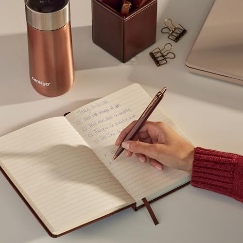 Eine Hand schreibt in ein Notizbuch. Eine Kupferflasche und ein Laptop sind in der Nähe.