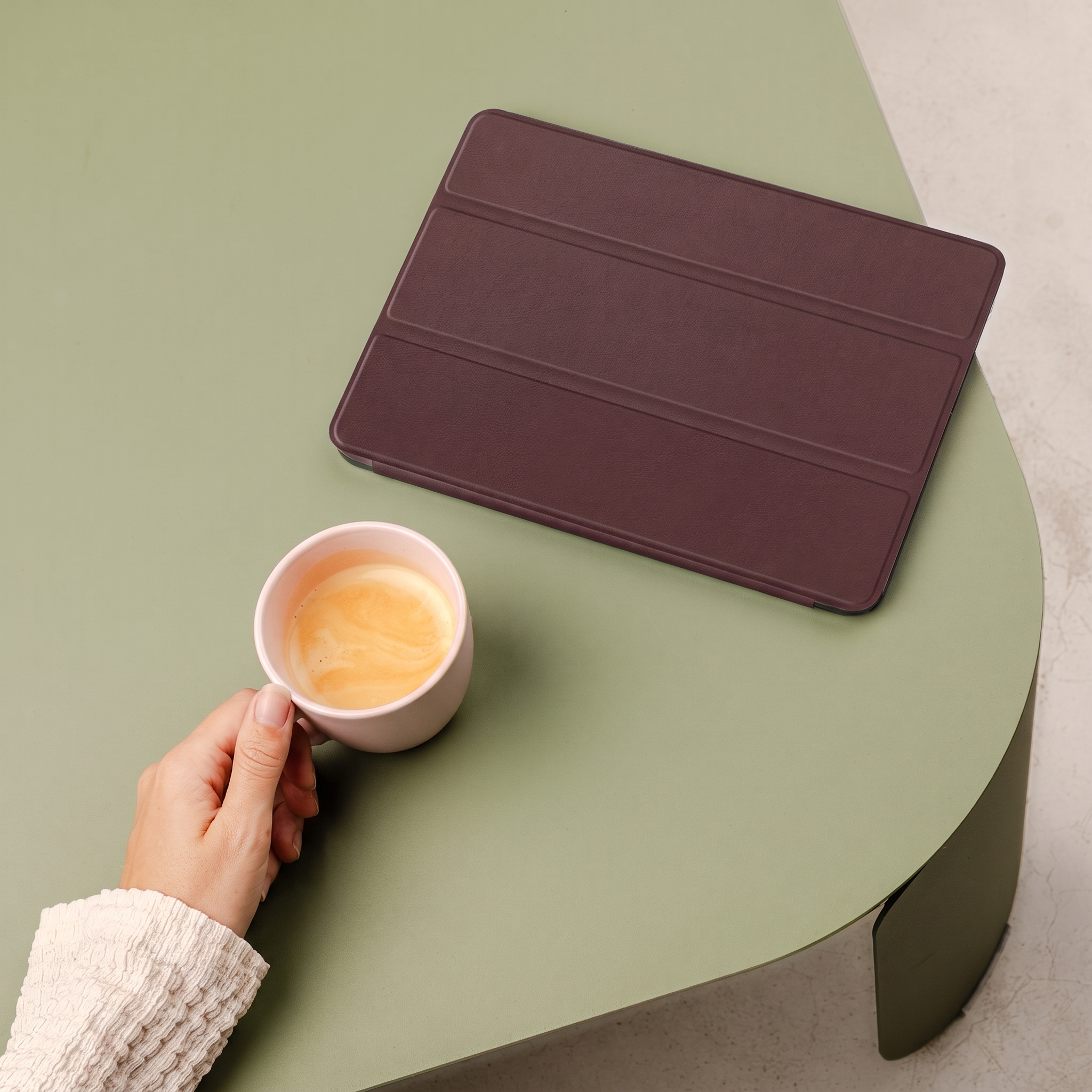 Une main tient une tasse rose de café à côté d'une tablette marron sur une table verte.