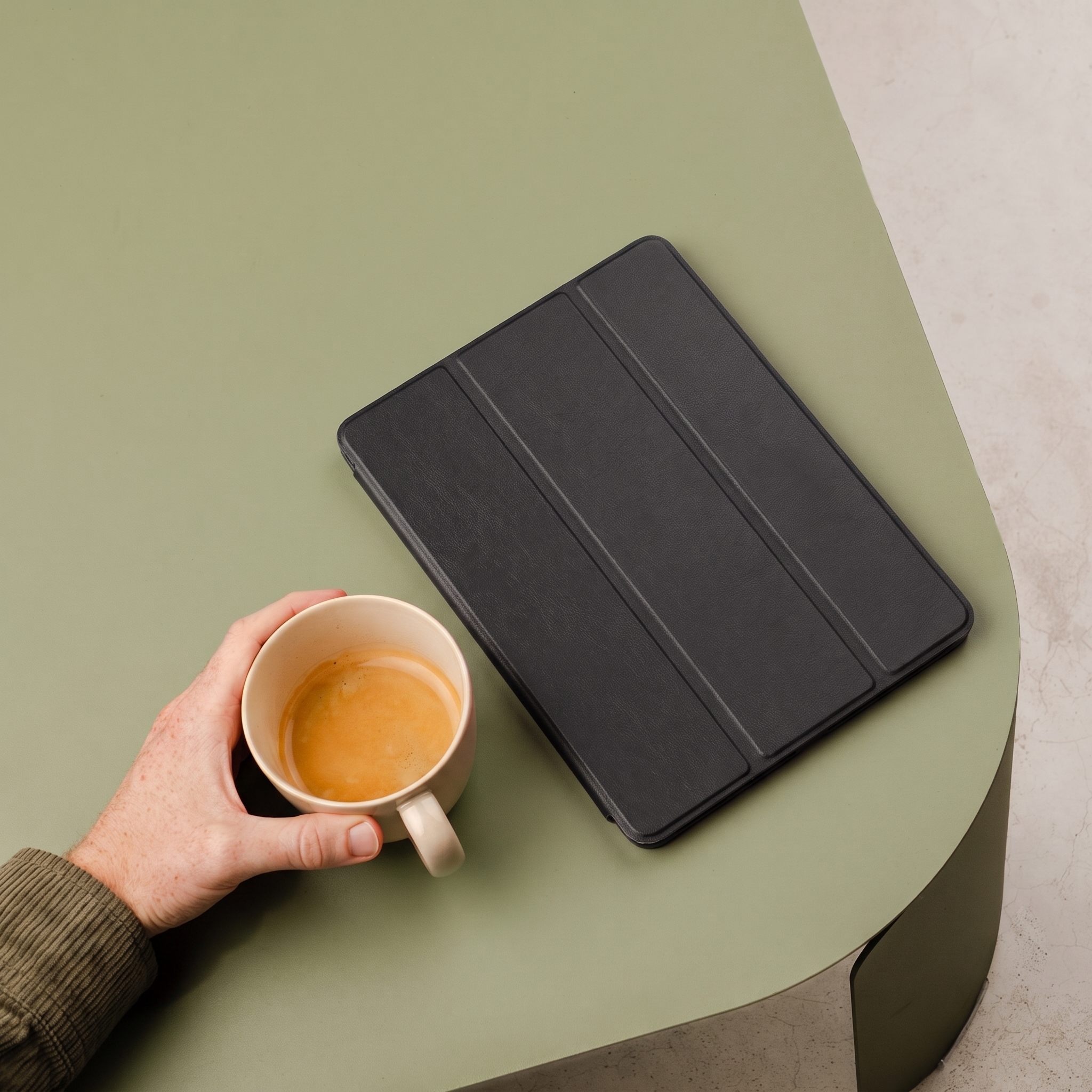 A hand holding a coffee cup next to a black tablet on a green table.