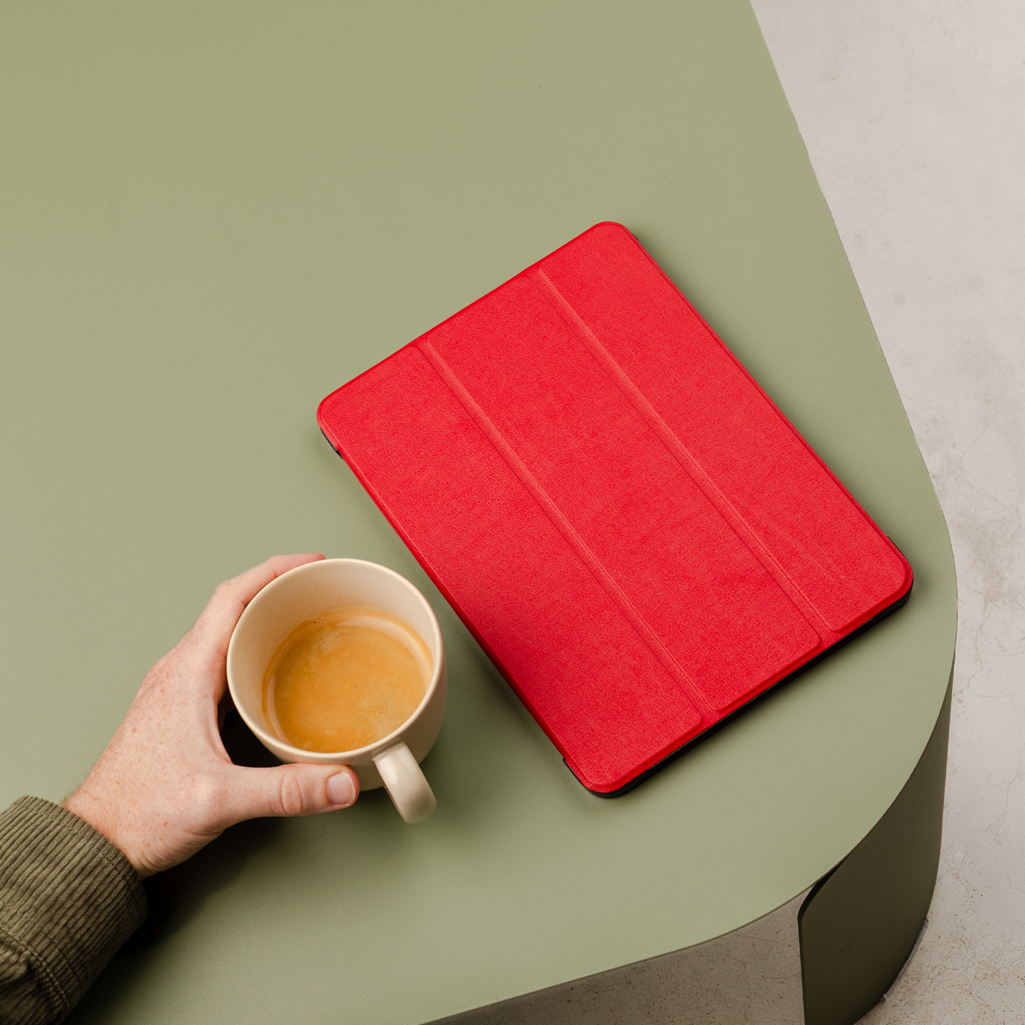 Una mano tiene una tazza di caffè accanto a una custodia rossa per tablet su un tavolo.