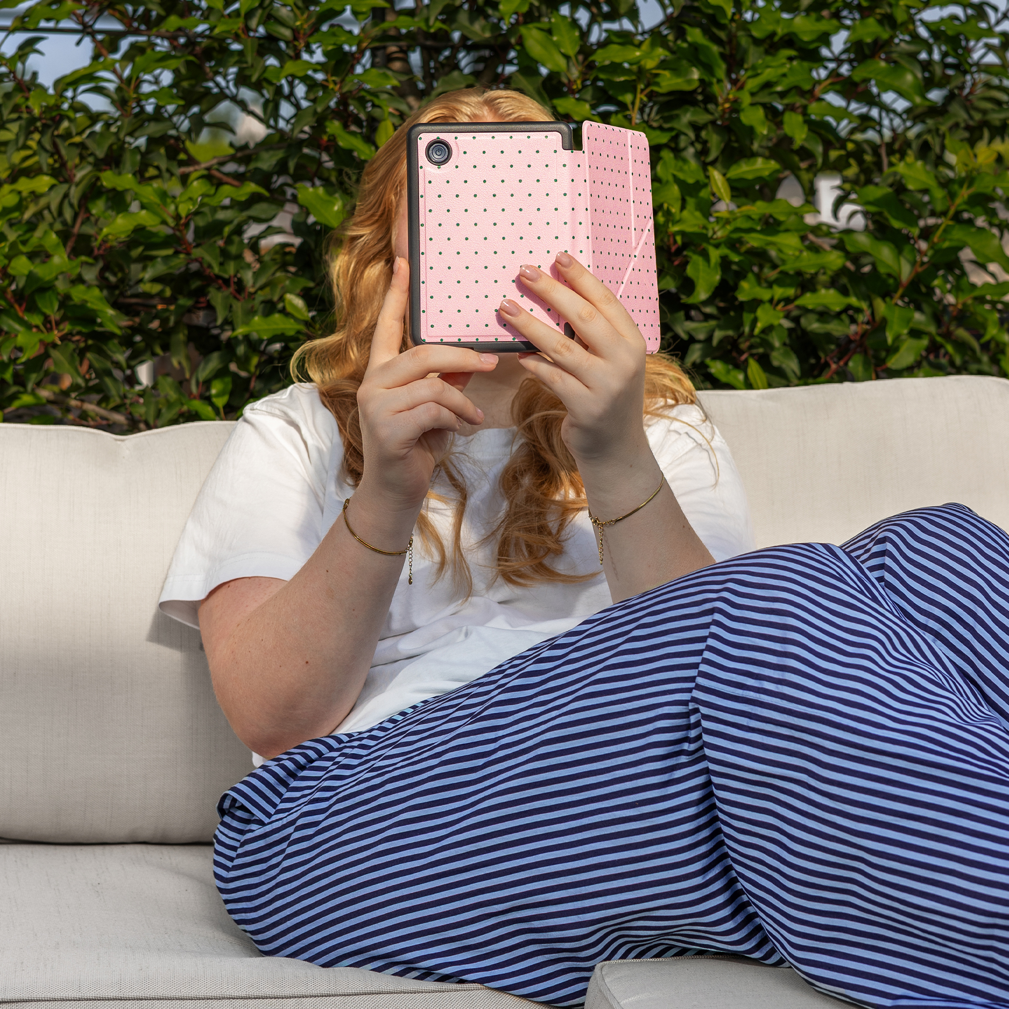 A person with red hair sits outdoors holding a pink, polka-dotted tablet.