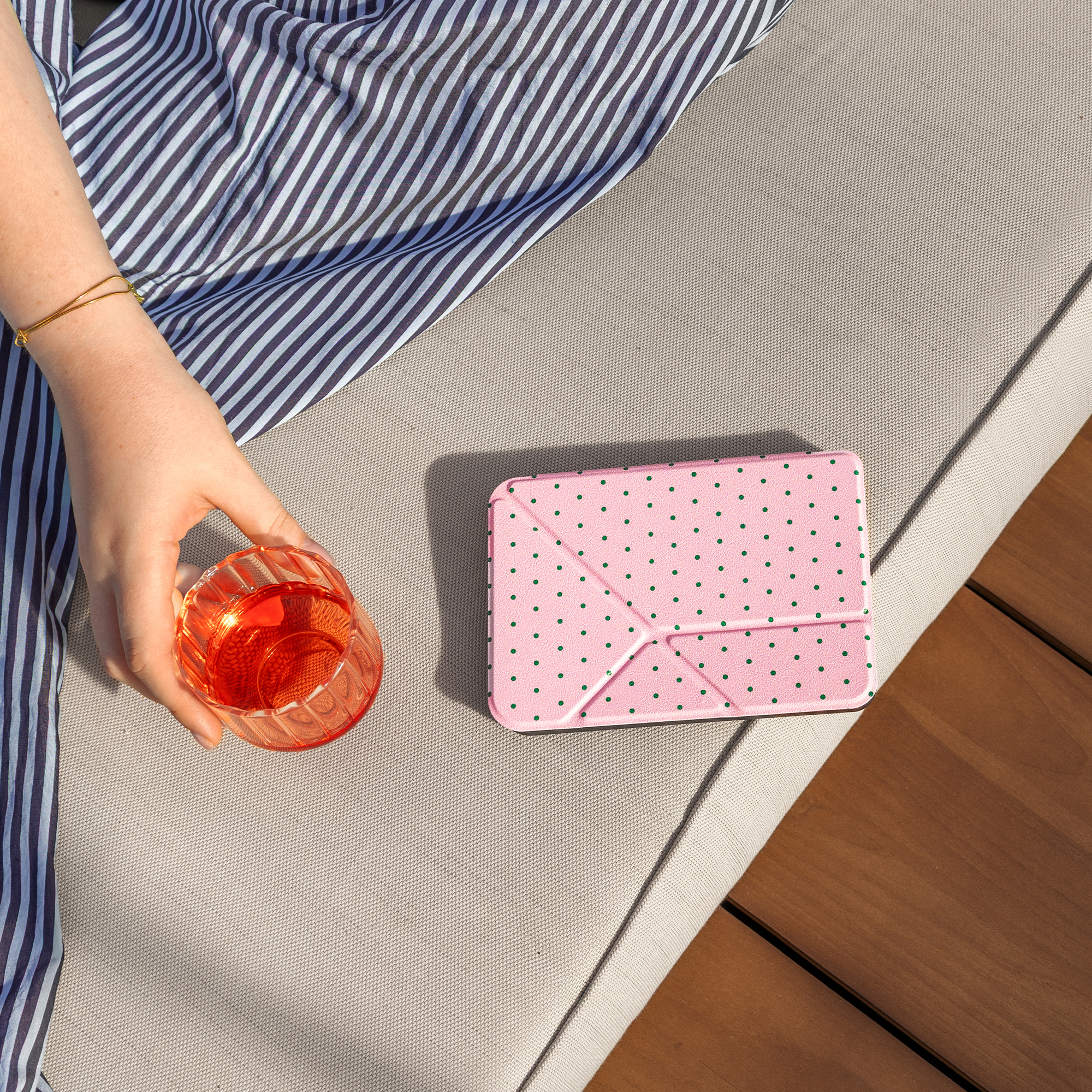 A hand holds a glass of red drink next to a pink case with green dots.