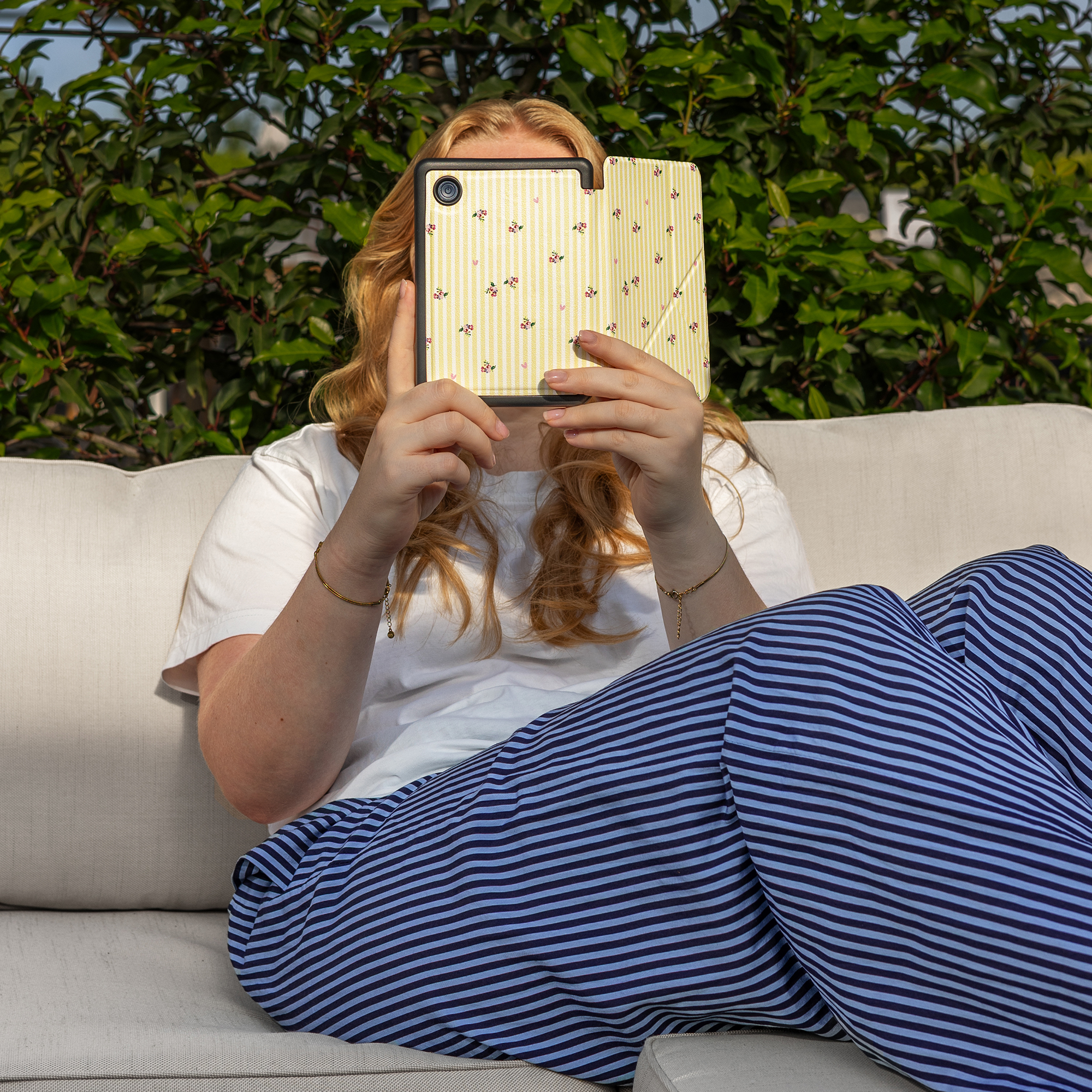 Vrouw met blond haar zit op een bank en houdt een tablet met bloemenpatroon vast.
