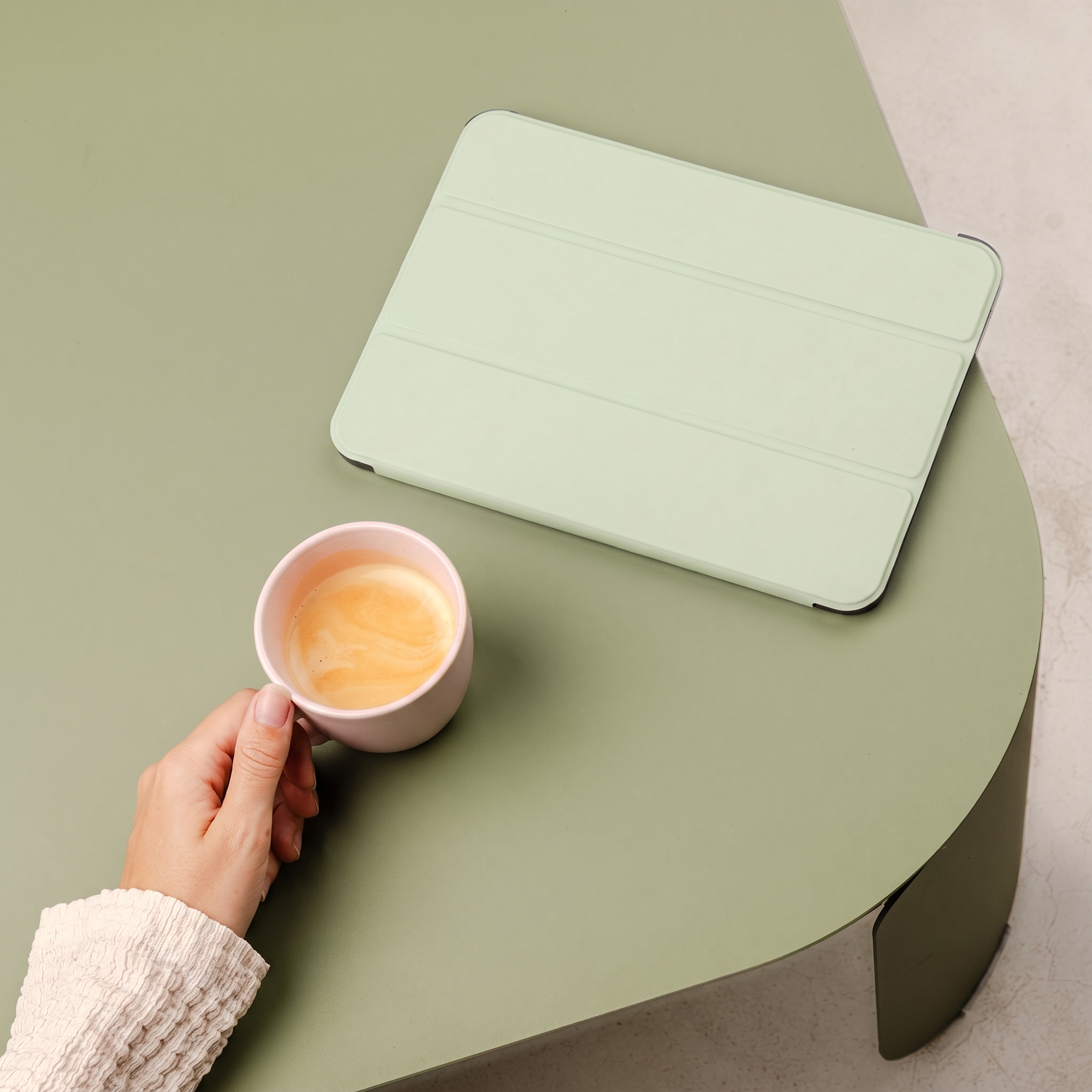 A hand holds a pink cup of coffee on a green table next to a tablet.