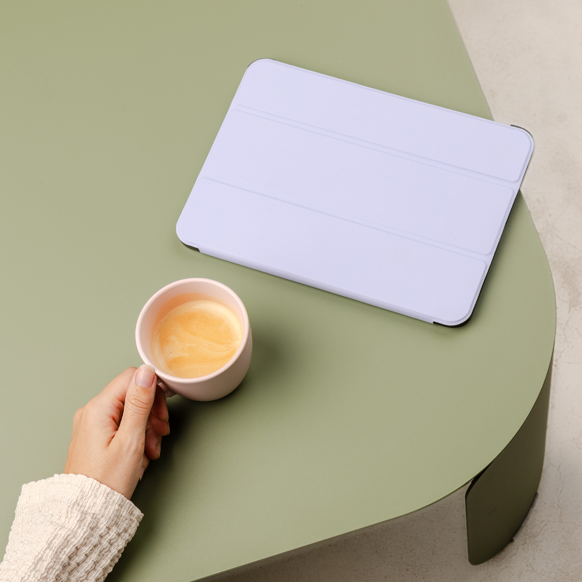 Una mano tiene una tazza di caffè rosa accanto a un tablet viola su un tavolo verde.
