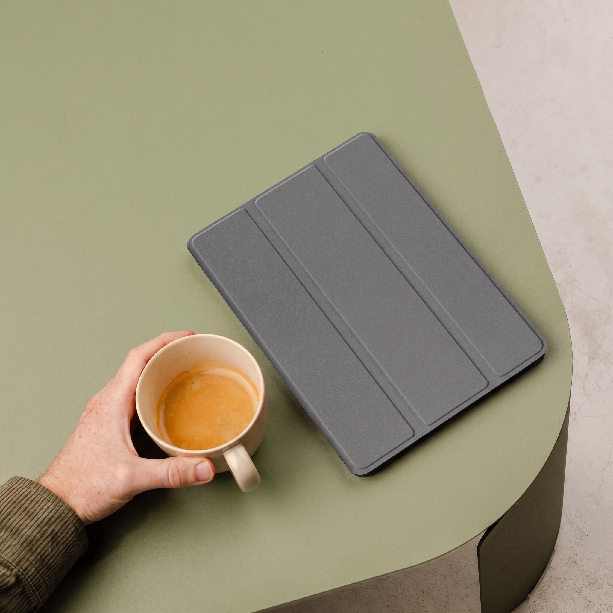 A hand holds a coffee cup next to a gray tablet on a green table.