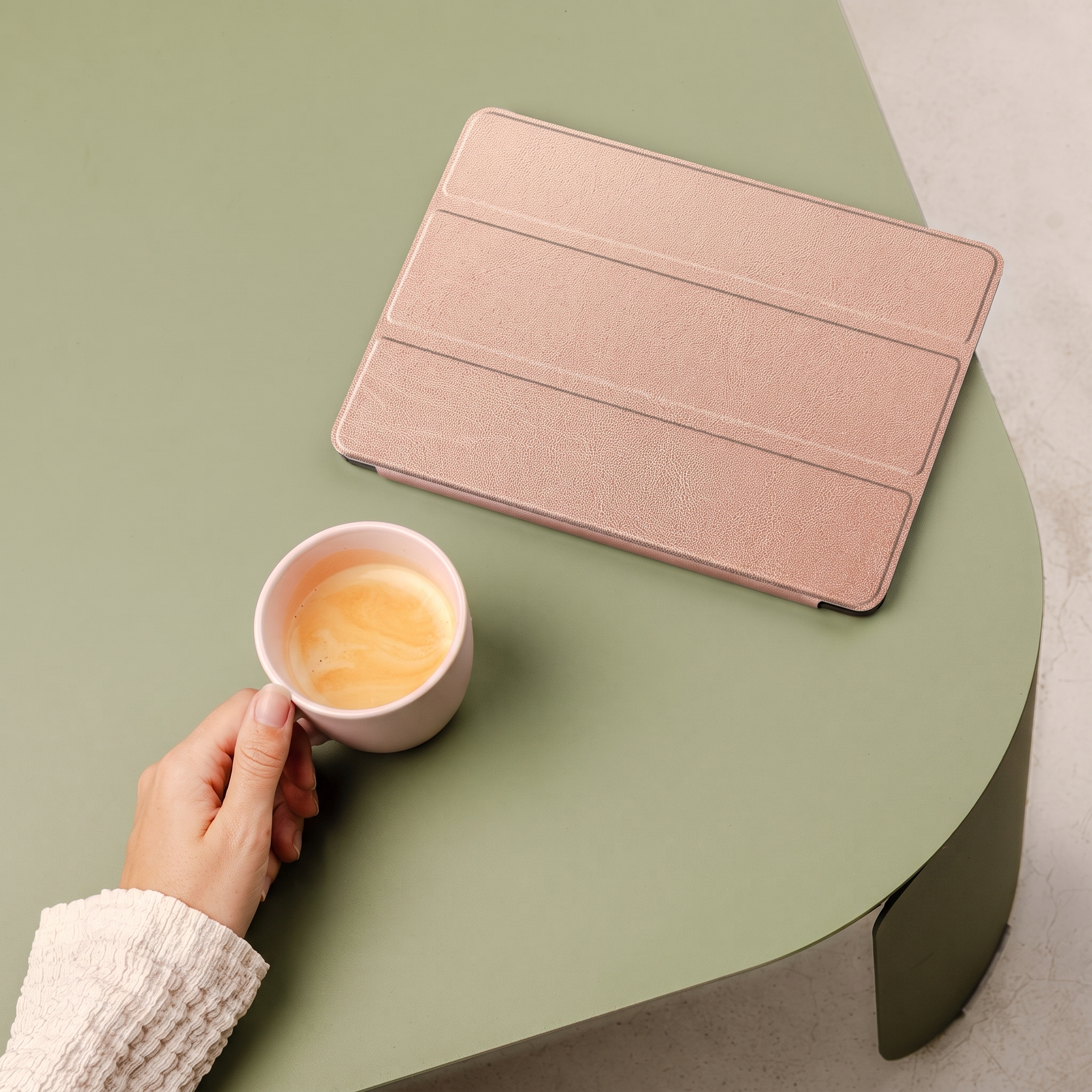 A hand holds a pink cup of coffee next to a rose gold tablet case on a green table.