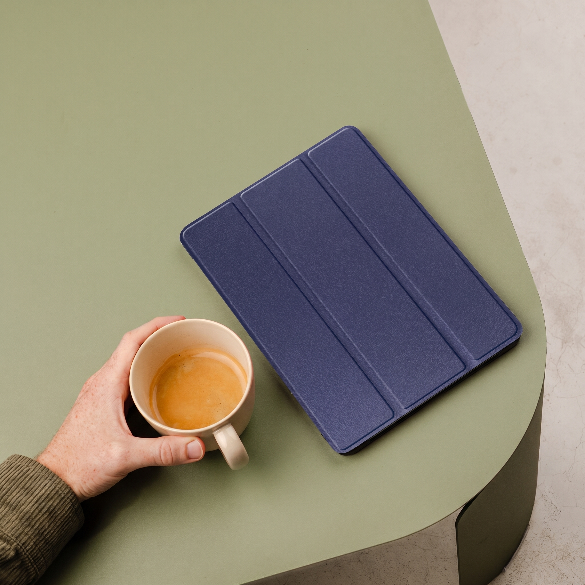 Une main tient une tasse de café à côté d'une housse de tablette bleue sur une table.
