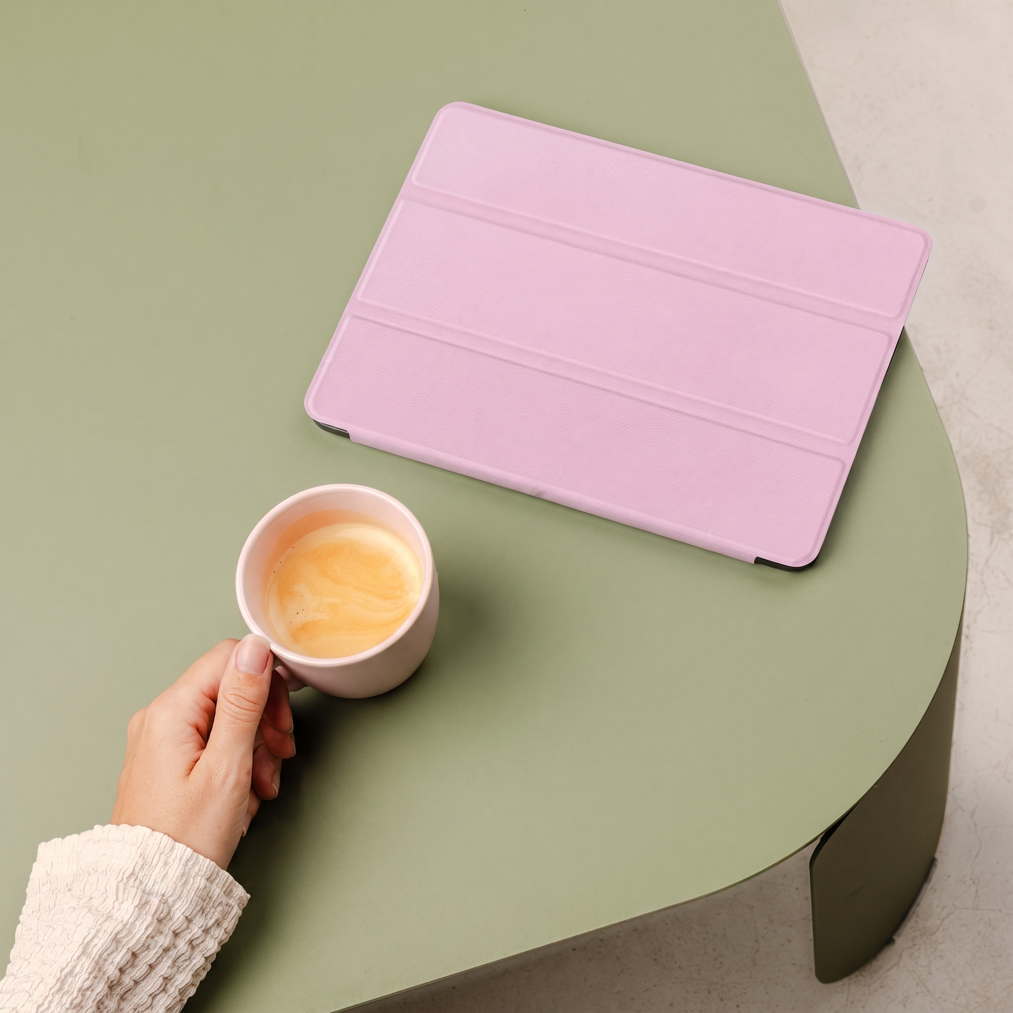 Une main tient une tasse de café à côté d'un support de tablette rose sur une table verte.