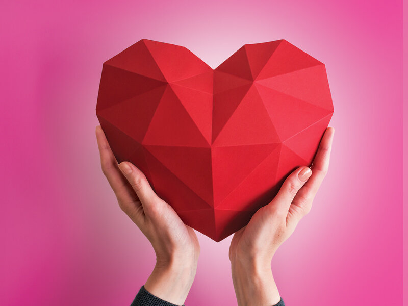 Dos manos sostienen un corazón rojo facetado sobre un fondo rosa.